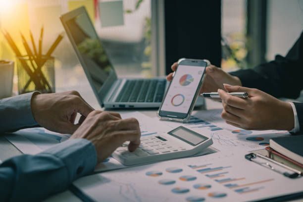 Two businessmen with pens pointing at financial market analysis charts using laptops while analyzing documents in hand with financial concept calculator.